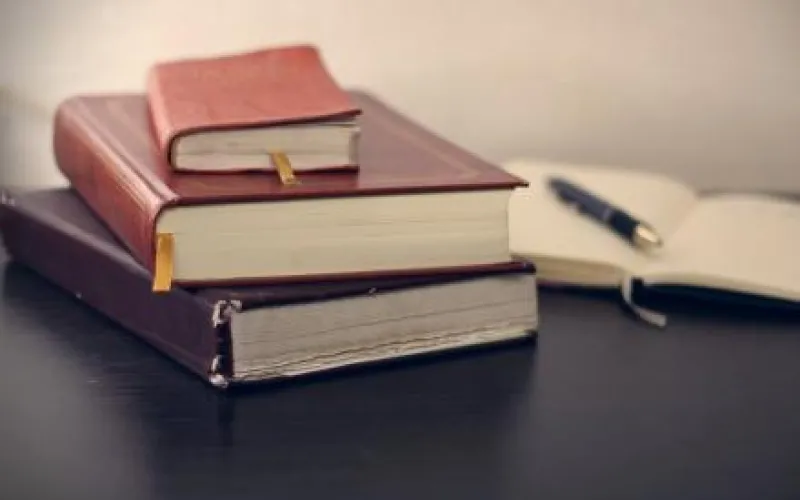 Stack 3 books, each smaller than the one below. There is an open notebook besides the stack, with a pen on top of the open page.