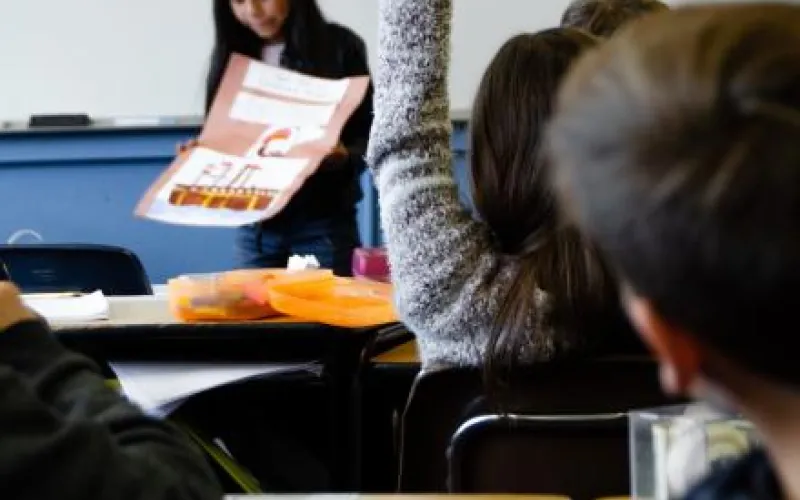 View from the end of a room, with a girl in the front raising her hand. Another women has a large chart in hand and seems to be placing it on the desk.