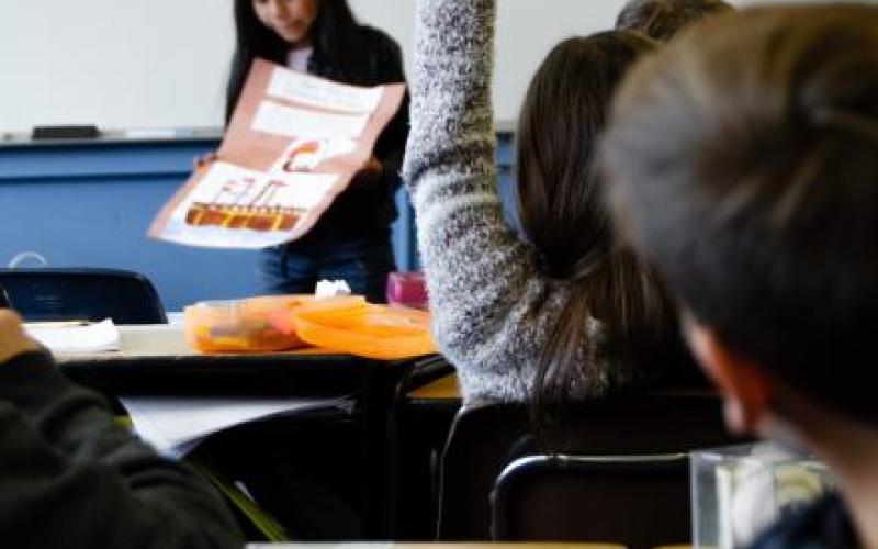 View from the end of a room, with a girl in the front raising her hand. Another women has a large chart in hand and seems to be placing it on the desk.