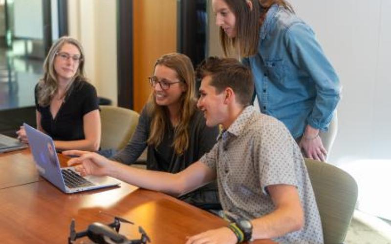 A group of 3 girls and one boy focused on a laptop screen, with the boy pointing to something at the laptop screen.